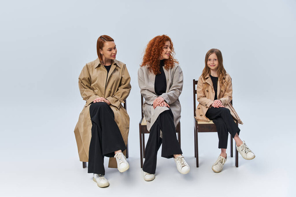 trzy pokolenia rudowłosych kobiet w płaszczach siedzących na krzesłach i patrzących na dziewczynę na szarym tle - Zdjęcie, obraz
