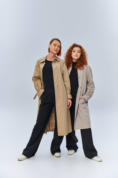 two generations of redhead women posing in autumnal coats on grey background, hands in pockets - Photo, Image