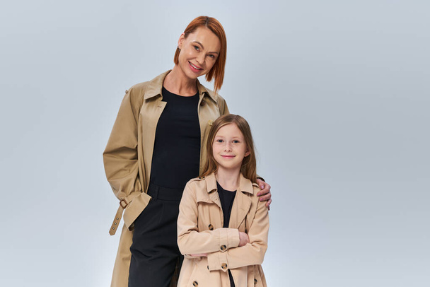 two generations, woman with red hair and little girl standing in trench coats on grey background - Photo, Image