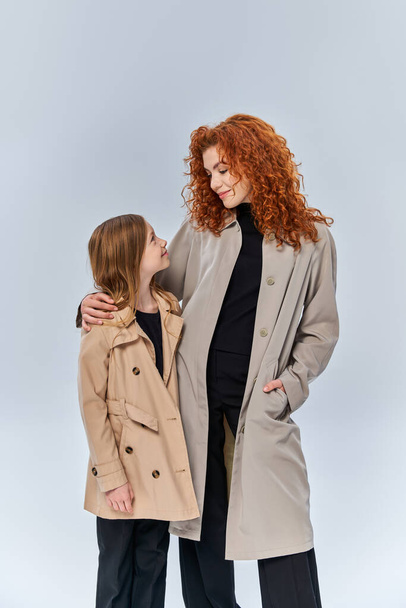 twee vrouwelijke generaties, roodharige vrouw kijken naar dochter en staan in jassen op grijze achtergrond - Foto, afbeelding