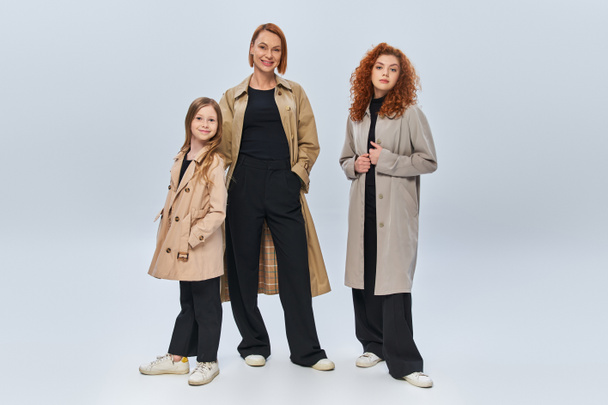 Trençkotlu mutlu kızıl saçlı aile gri arka planda birlikte poz veriyor, üç kadın jenerasyon. - Fotoğraf, Görsel