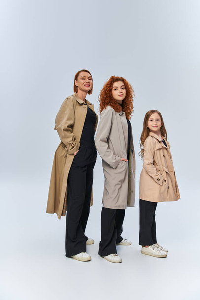 joyful redhead family in coats posing with hands in pockets on grey background, female generations - Photo, Image