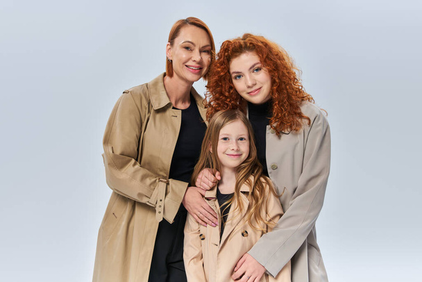 redhead women in autumn coats hugging little girl on grey background, three female generations - Photo, Image
