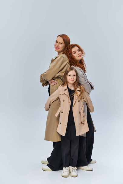 redhead girl hugging happy relatives in coats on grey background, female generations, full length - 写真・画像