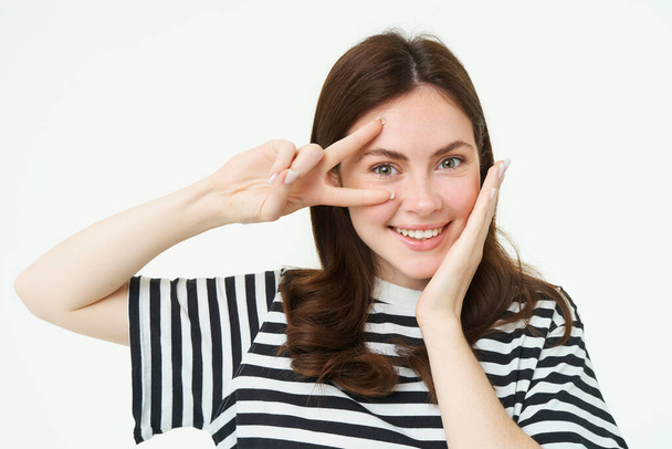 Portrait of beautiful, smiling female model, touches her clear glowing skin, shows peace sign and looks happy at camera, isolate on white background. - Photo, Image