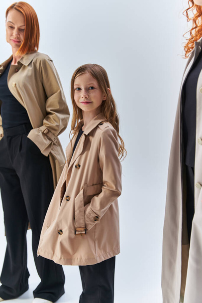 chica con abrigo posando con las manos en los bolsillos cerca de la familia pelirroja, tres generaciones de concepto de mujer - Foto, Imagen