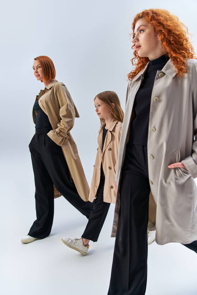 three generation redhead family walking with hands in pockets of trench coats on grey backdrop - Photo, Image
