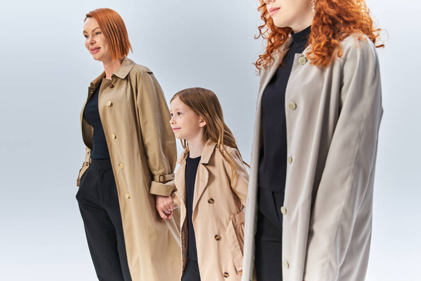 trzypokoleniowa rodzina rudowłosych spacerująca razem w stylowych płaszczach na szarym tle, jesienna moda - Zdjęcie, obraz