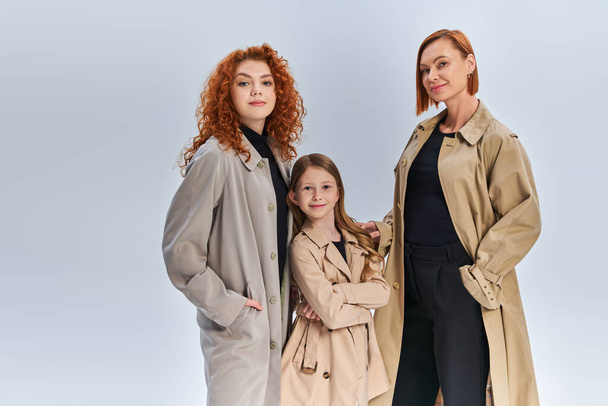 three generation joyful family standing together in stylish coats on grey backdrop, autumn fashion - Фото, изображение