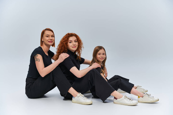 Drei Generationen Konzept, fröhliche rothaarige Familie sitzt in passenden Outfits vor grauem Hintergrund - Foto, Bild