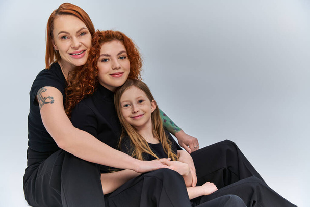concetto di tre generazioni, famiglia rossa in abiti coordinati che si abbracciano e si siedono su sfondo grigio - Foto, immagini