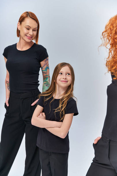 redhead girl standing with folded arms near women in matching clothes on grey backdrop, generations - Photo, Image