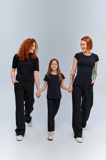 cheerful girl holding hands and walking with redhead family in matching outfits on grey backdrop - Photo, Image