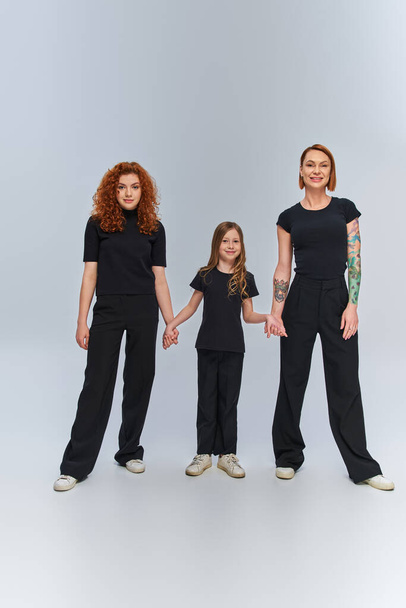 cheerful girl holding hands and standing with redhead women in matching outfits on grey backdrop - Photo, Image
