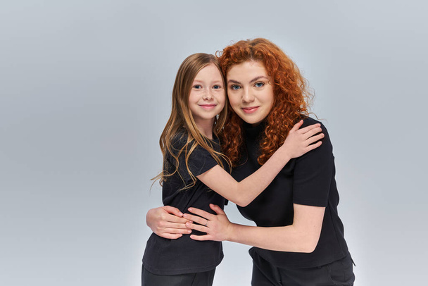 happy young mother with curly red hair hugging little girl on grey backdrop, two generations - Photo, Image