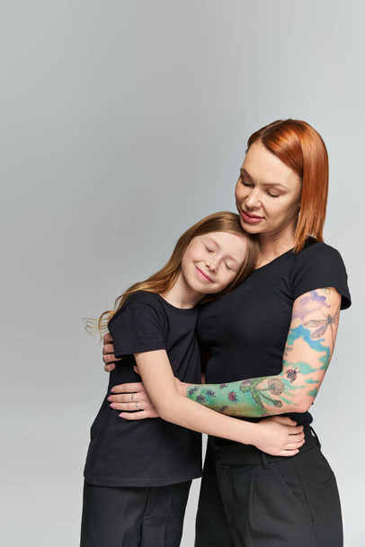 два поколения, рыжеволосая женщина и девочка в чердаке, обнимающиеся на сером заднем плане, семейная любовь - Фото, изображение