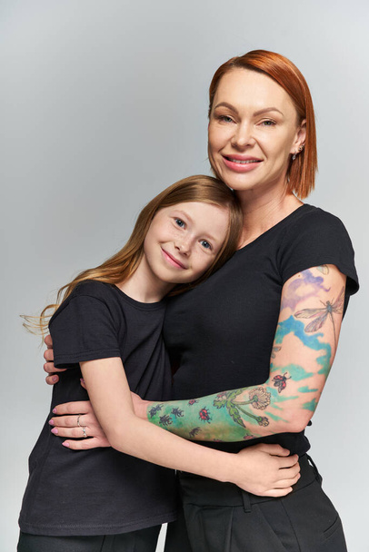two generations, redhead woman and girl in matching attire hugging on grey backdrop, happiness - Foto, Bild