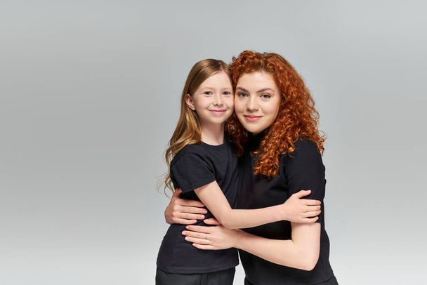 two generations concept, happy redhead mother and child in matching attire hugging on grey backdrop - Zdjęcie, obraz