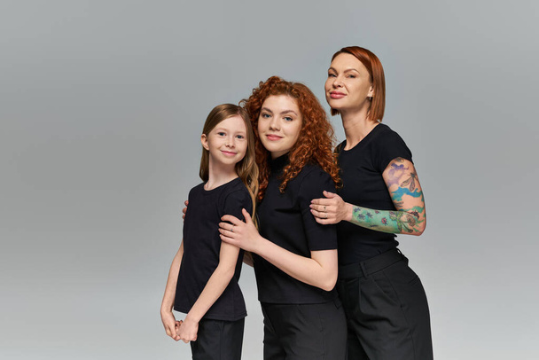 концепція жіночих поколінь, щасливі руді жінки і дитина в відповідному вбранні, обіймаючись на сірому тлі - Фото, зображення