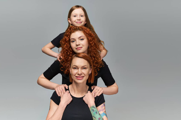 weibliches Generationen-Konzept, glückliche rothaarige Familie in passender Kleidung, die sich auf grauem Hintergrund umarmt - Foto, Bild