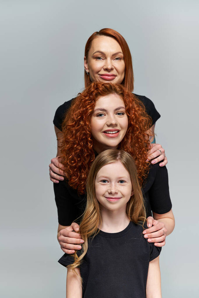 concepto de generaciones, familia sonriente con el pelo rojo posando en trajes a juego sobre fondo gris - Foto, imagen