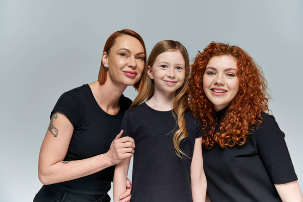 family bond, redhead women and freckled girl in matching attire smiling on grey background - Foto, imagen