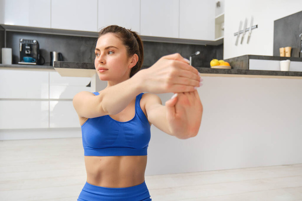 Portrait of beautiful woman does fitness training, sits on floor near kitchen, stretching her hands, workout from home. Copy space - Photo, Image