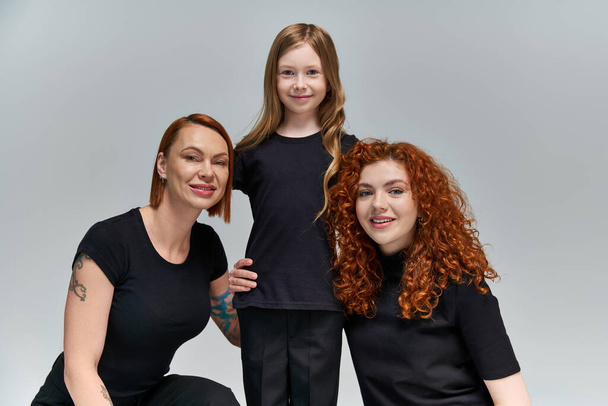 family portrait, happy freckled girl hugging redhead women in matching clothes on grey background - Photo, Image