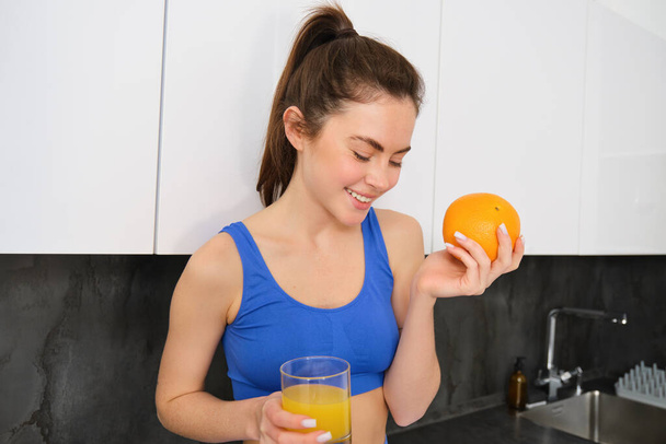 Welzijn en sport. Jonge lachende voedingsdeskundige, fitness meisje met sinaasappel en vers sap, drinken uit glas en er gelukkig uitzien, poseren in sportbeha en leggings, staan in de keuken. - Foto, afbeelding