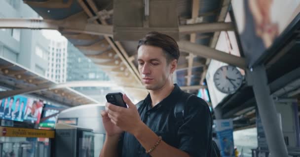 Jongeman gebruiken smartphone in metrostation in afwachting van aankomst komende metro trein. 30s man passagier in het openbaar vervoer te stoppen wacht op de tram in de moderne stad. Vervoer met technologie. - Video