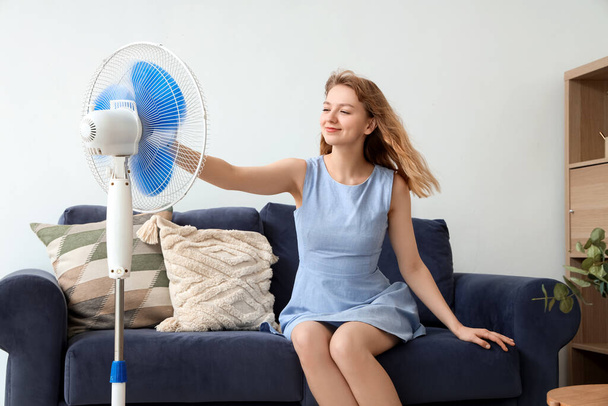 Щаслива красива молода жінка з електричним вентилятором сидить на дивані у вітальні - Фото, зображення