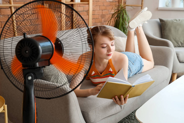 Молодая женщина с дующим электрическим вентилятором читает книгу на диване дома - Фото, изображение