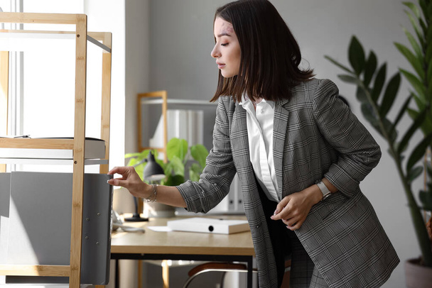 Jonge zakenvrouw neemt map van plank in kantoor - Foto, afbeelding