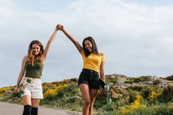 Счастливые многоэтнические подруги, держащиеся за руки во время прогулки по дороге с длинными досками - Фото, изображение