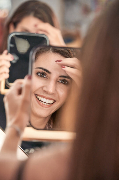 Радостный урожай женщины с длинными волосами, глядя на отражение в зеркале с улыбкой, сидя в современном светлом зеркале во время встречи - Фото, изображение