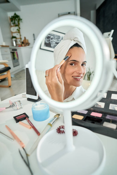 Wysoki kąt odbicia w lustrze uśmiechniętej kobiety z ręcznikiem na głowie nakładającym podkład na twarz pędzlem podczas codziennego makijażu w domu w godzinach porannych - Zdjęcie, obraz