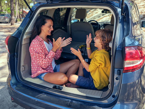 Позитивная мать с сыном, играющая в игру с тортом и смотрящая друг на друга, сидя в грузовике автомобиля, припаркованного в сельской местности в летний день - Фото, изображение