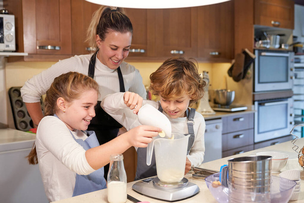 Delighted young mother in casual clothes and apron standing behind cute son and daughter weighting flour while helping to bake in kitchen - Photo, Image