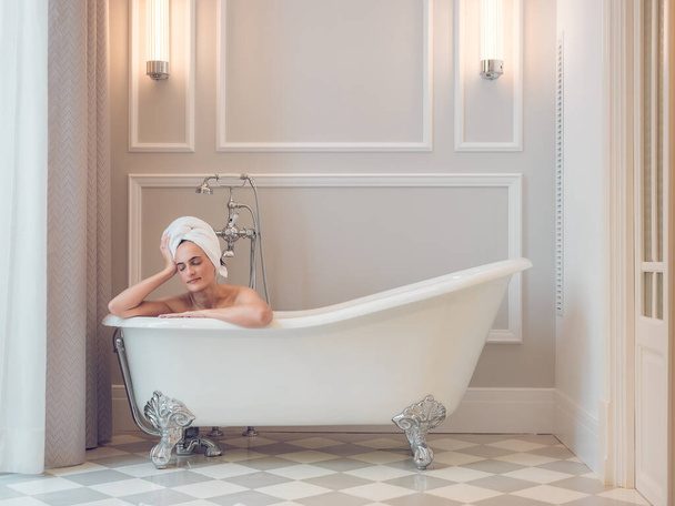 Prachtige vrouw met handdoek op het hoofd zitten in witte badkuip in licht klassieke badkamer overdag - Foto, afbeelding