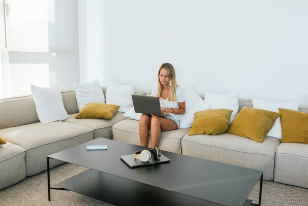自宅の軽いリビングルームでクッションで快適なソファに座っている間,ネットブックにタイピングするカジュアルな着用に集中した女性 - 写真・画像
