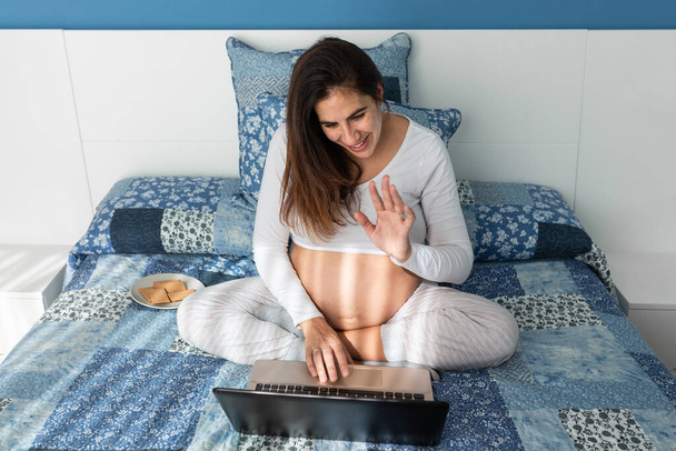 Από ψηλά χαμογελώντας έγκυος γυναίκα μιλώντας σε βίντεο κλήση στο netbook και χαιρετώντας το χέρι, ενώ κάθεται στο άνετο κρεβάτι στο σπίτι - Φωτογραφία, εικόνα