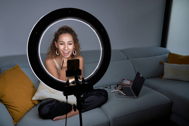 明るい女性ブロガーの完全な体はソファに座って、スマートフォンや照明リングランプ上のソーシャルネットワークのためのメイクチュートリアルビデオを撮影しながら、ブラシで口紅を適用します - 写真・画像