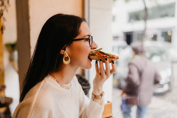 Вид на красивую молодую женщину в стильном наряде, кусающую вкусный сэндвич, сидя у окна в уютном кафе - Фото, изображение