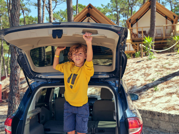 Child holding door of car trunk and looking at camera near cozy huts placed in forest on sunny day during summer vacation - Photo, Image