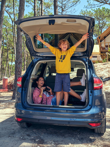 Pełna długość kobiety trzymającej lornetkę i patrzącej na podekscytowanego nastolatka stojącego w bagażniku samochodu w pobliżu glamping campingu w lesie i cieszącego się swobodą podczas wakacji letnich - Zdjęcie, obraz