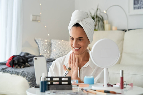 Positive smiling female blogger with towel on head showing cosmetic applicator while recording video for skincare routine blog at home near couch with sleeping dog - Photo, Image