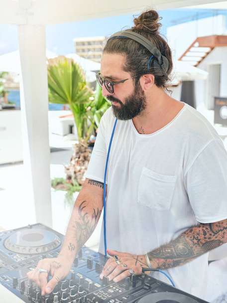 Focused bearded tattooed disc jockey working on DJ mixer while standing in outdoor party zone at summer terrace of resort hotel - Fotografie, Obrázek