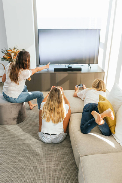 Πίσω όψη θετικών γυναικών φίλων με χειριστήρια παιχνιδιών που παίζουν βιντεοπαιχνίδι ενώ κάθονται στον καναπέ στο σαλόνι και στο έδαφος στο σπίτι - Φωτογραφία, εικόνα