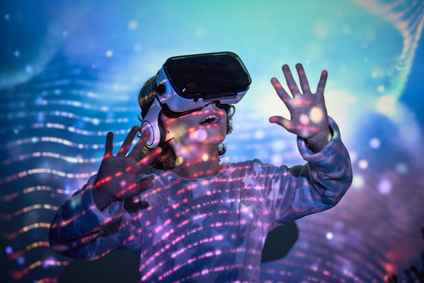 Verrukt jongetje in casual kleding en VR-headset glimlachen gelukkig en kijken omhoog tijdens het verkennen van de virtuele realiteit in de verlichte kamer - Foto, afbeelding