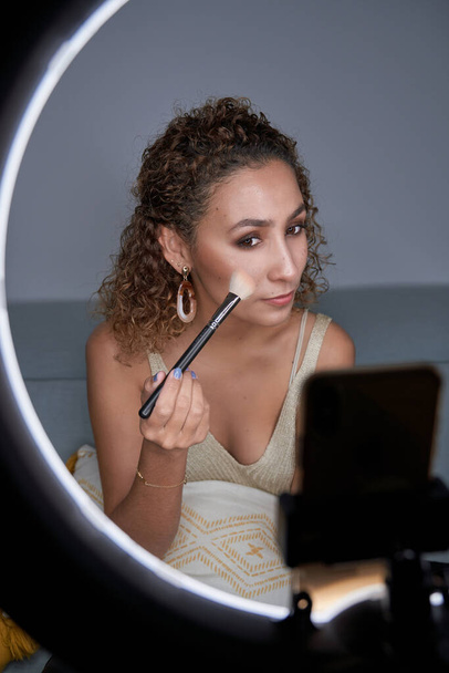 Mladý pozitivní krása blogger žena stínování zarudnutí s profesionálním štětcem na tváři při provádění on-line lekce make-upu přes mobilní telefon a pomocí osvětlené kruh lampy - Fotografie, Obrázek